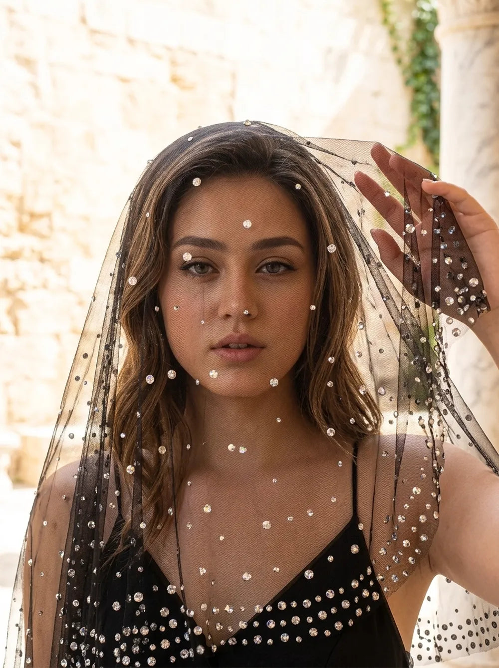 Front view of model holding up sheer black rhinestone veil frames around face with mysterious gothic allure