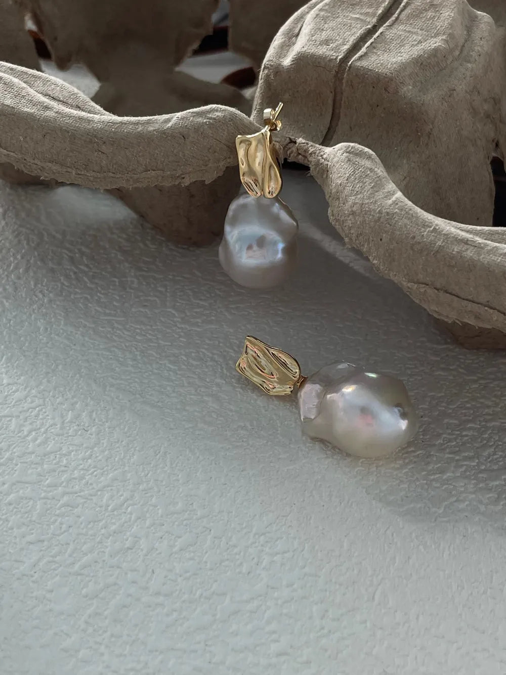 Molten metal baroque pearl drop earrings resting on textured paper pulp props.