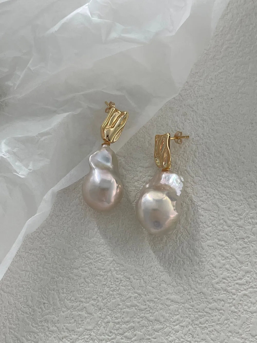 Molten metal baroque pearl stud earrings displayed on crinkled white paper background.
