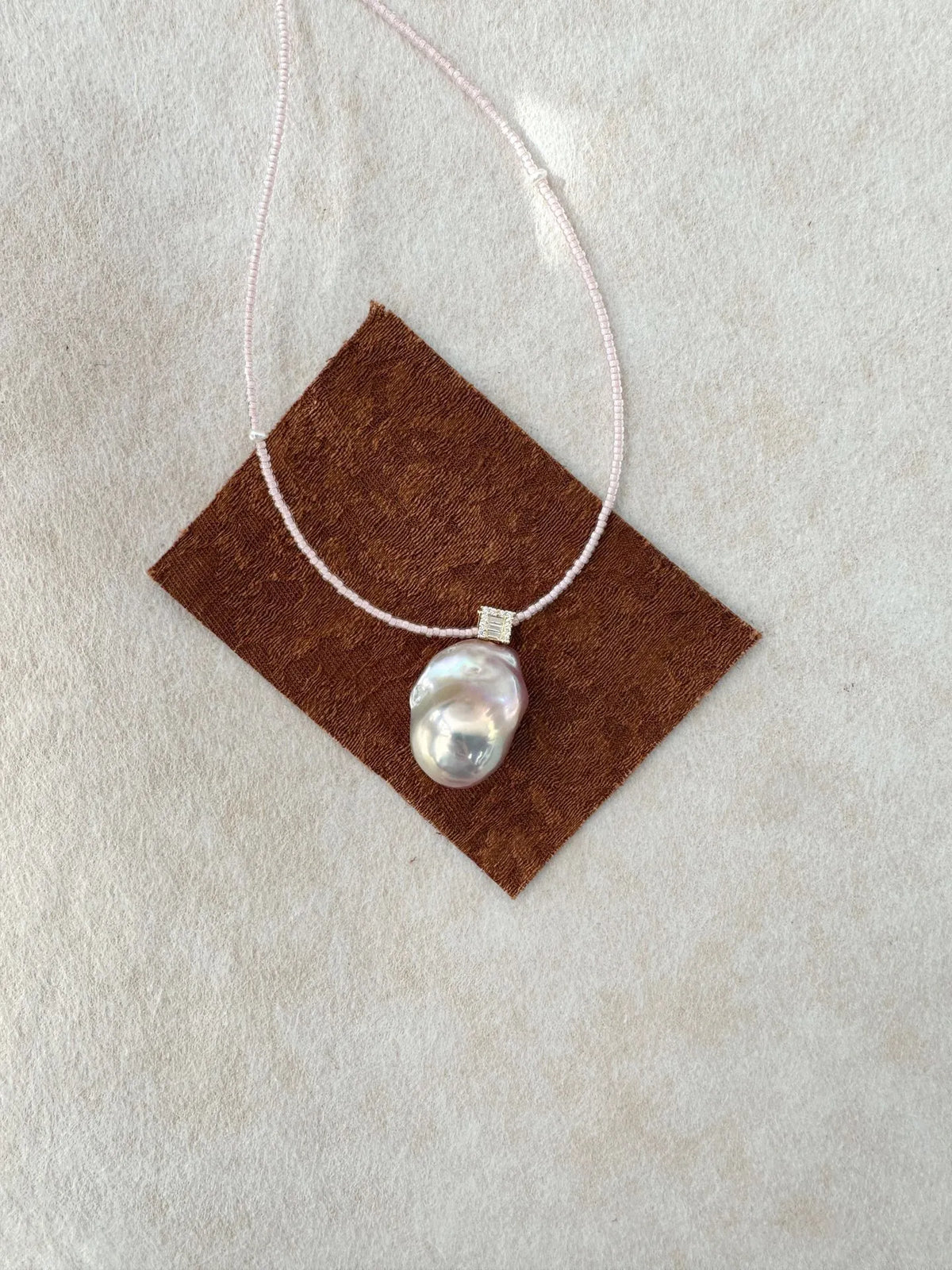 Baroque pearl pendant with pavé bail displayed on brown suede fabric.
