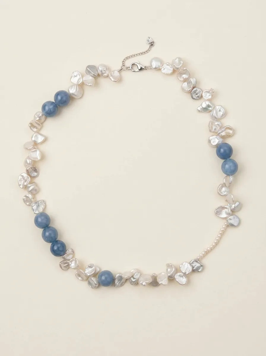 Full view flatlay of blue gemstone and petal baroque pearl choker on off-white background.