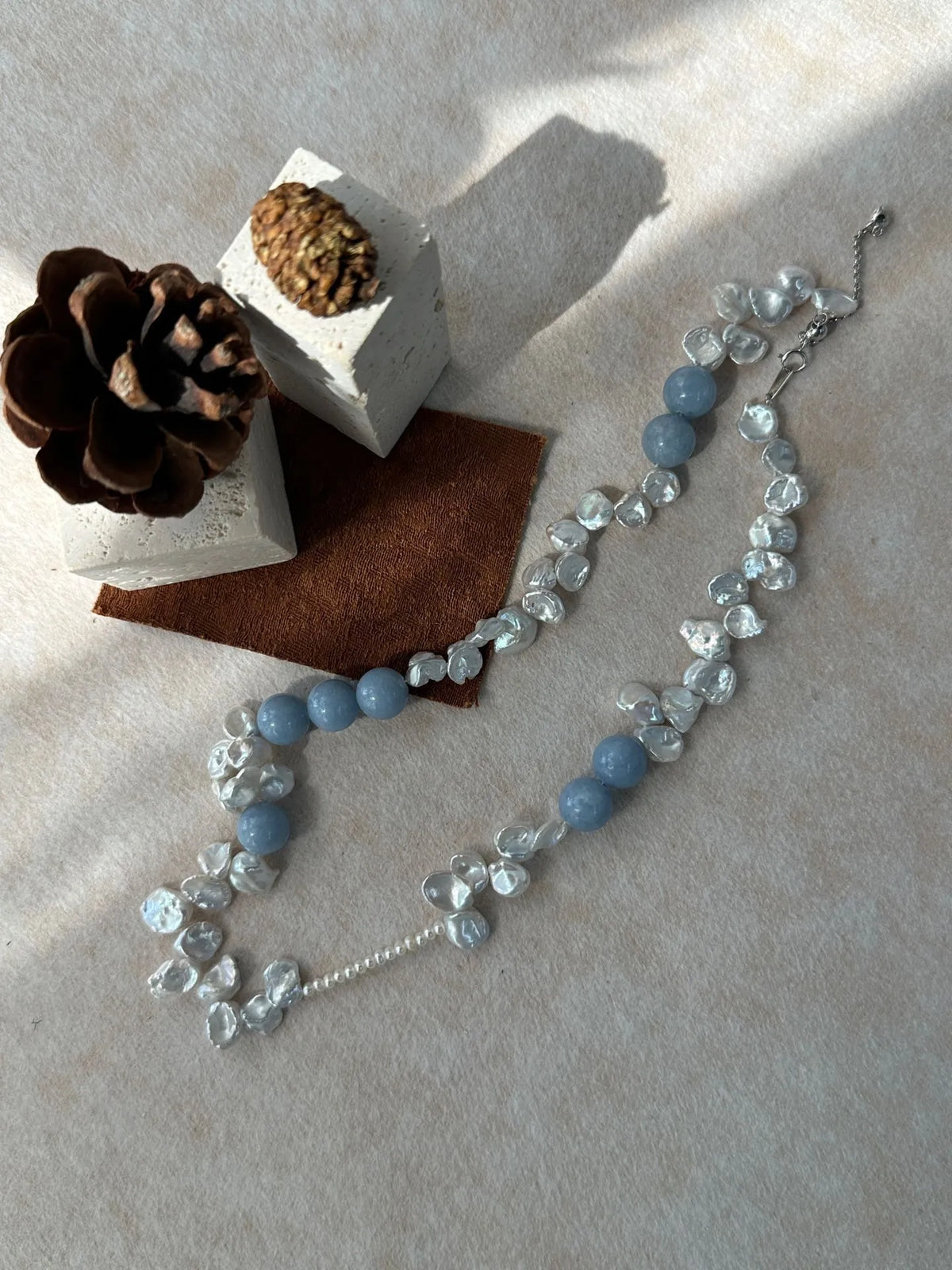 Blue gemstone and petal pearl necklace on beige fabric with brown velvet accent and pinecone prop.