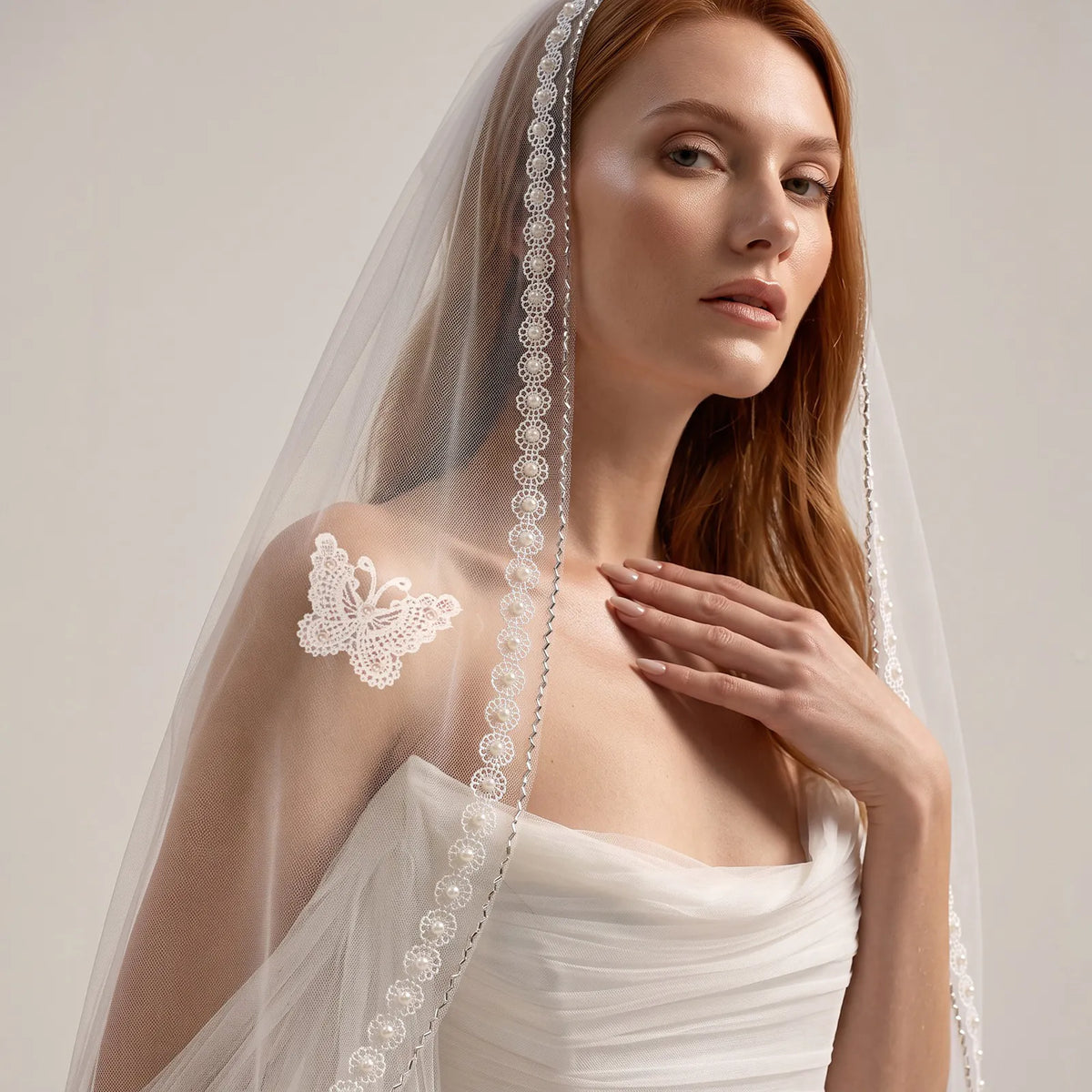 Front half-length portrait of red-haired bride wearing sheer tulle veil with butterfly lace applique near shoulder and detailed pearl trim