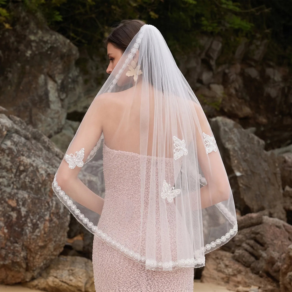 Rear profile of bride outdoors wearing 90cm fingertip veil over strapless blush dress, showing butterfly motifs against stone background
