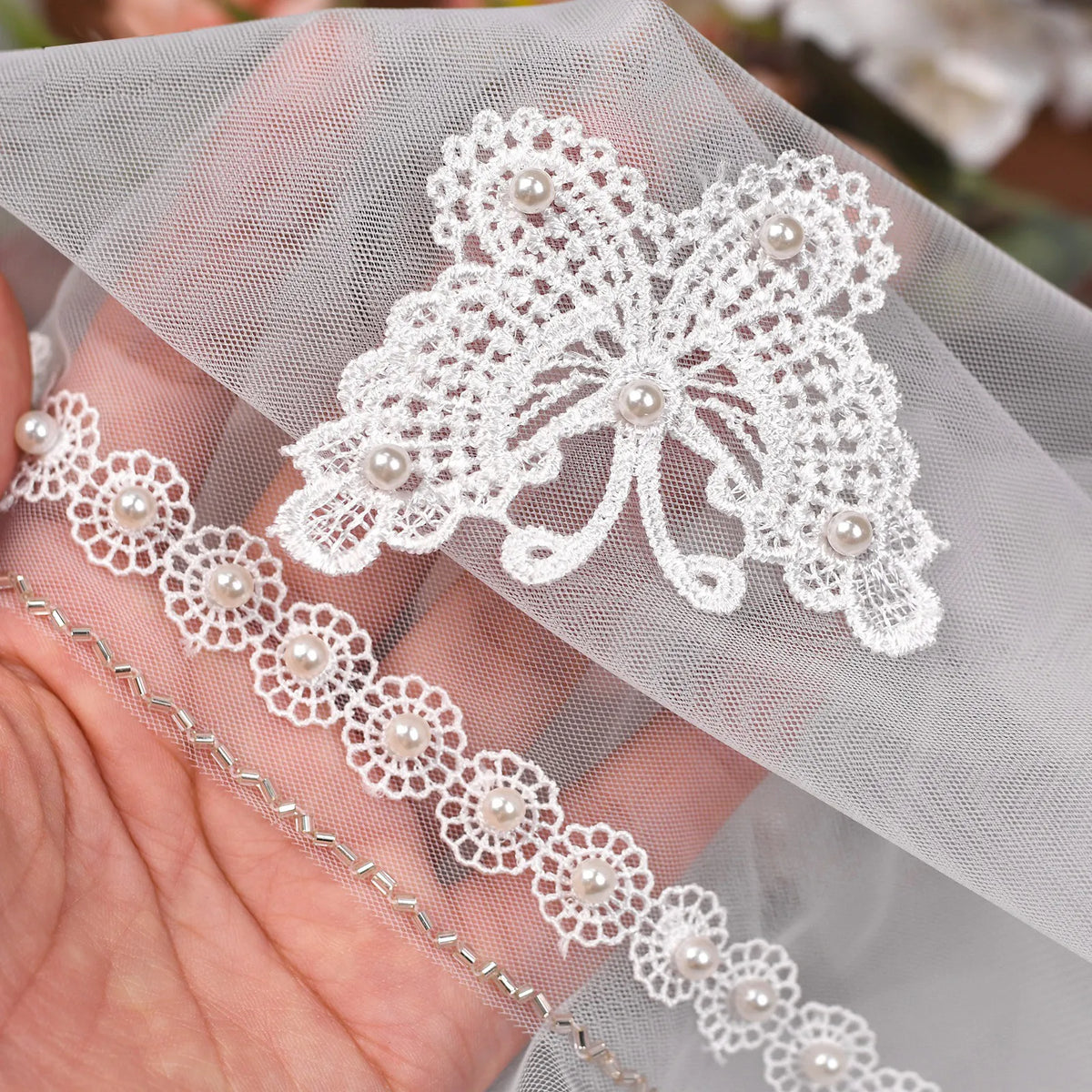 Hand holding veil tulle showing high-definition macro detail of embroidered 3D butterfly applique and delicate pearl beaded trim