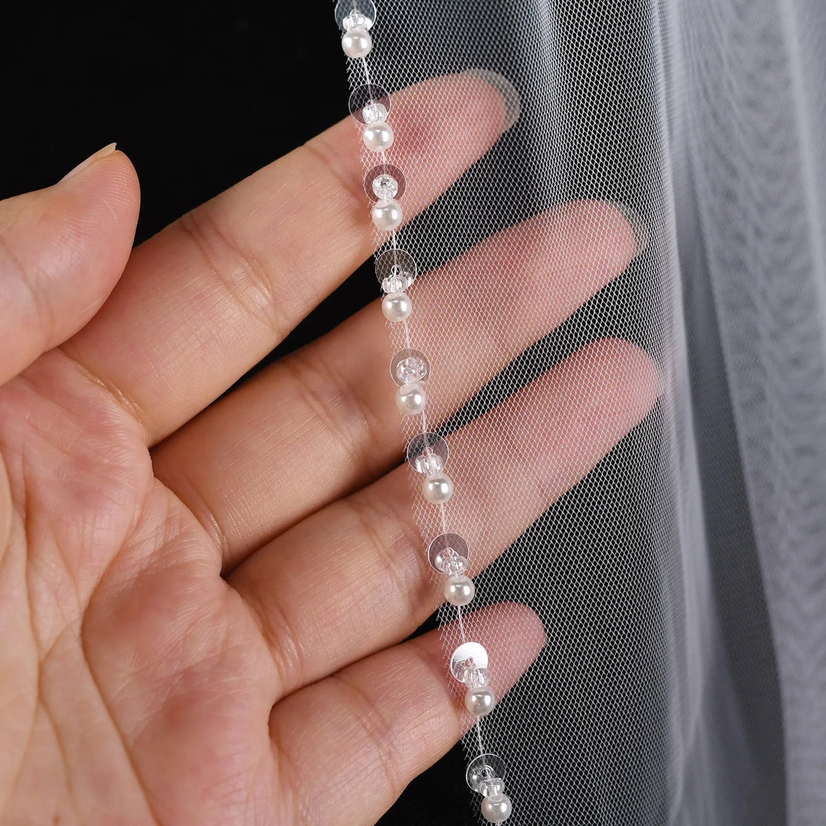 Extreme macro close-up of hand holding veil edge, showing delicate clear sequins, round white faux pearls, and high-transparency mesh net