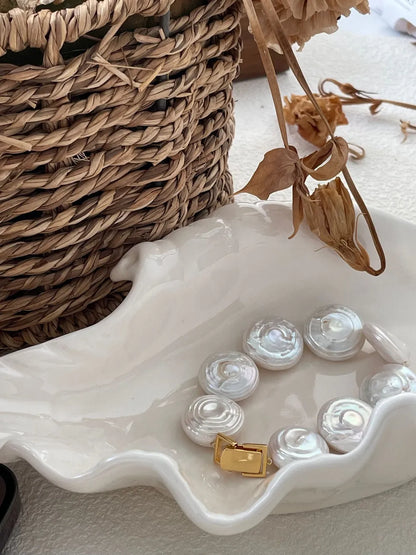 Coin baroque pearl bracelet resting inside a shell-shaped ceramic dish next to dried flowers.
