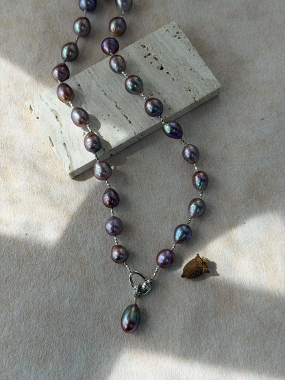 Deep violet baroque pear.l necklace displayed on textured stone board highlighting silver front ring and drop pendant.