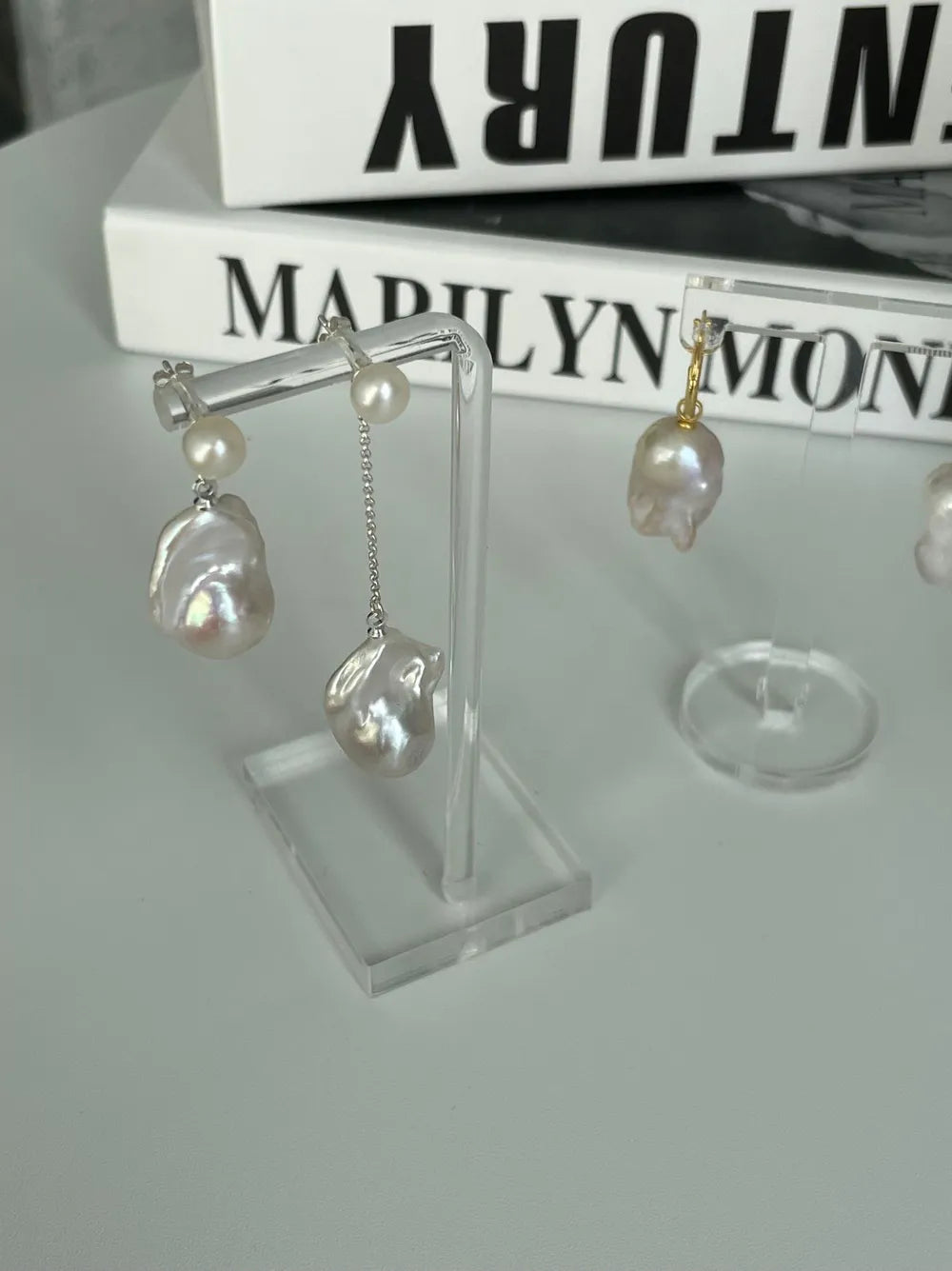 Flame-tailed baroque pearl drop earrings displayed on transparent acrylic stands.