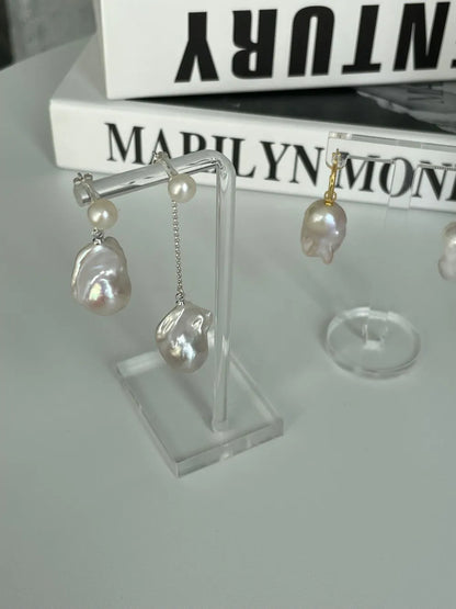 Flame-tailed baroque pearl drop earrings displayed on transparent acrylic stands.