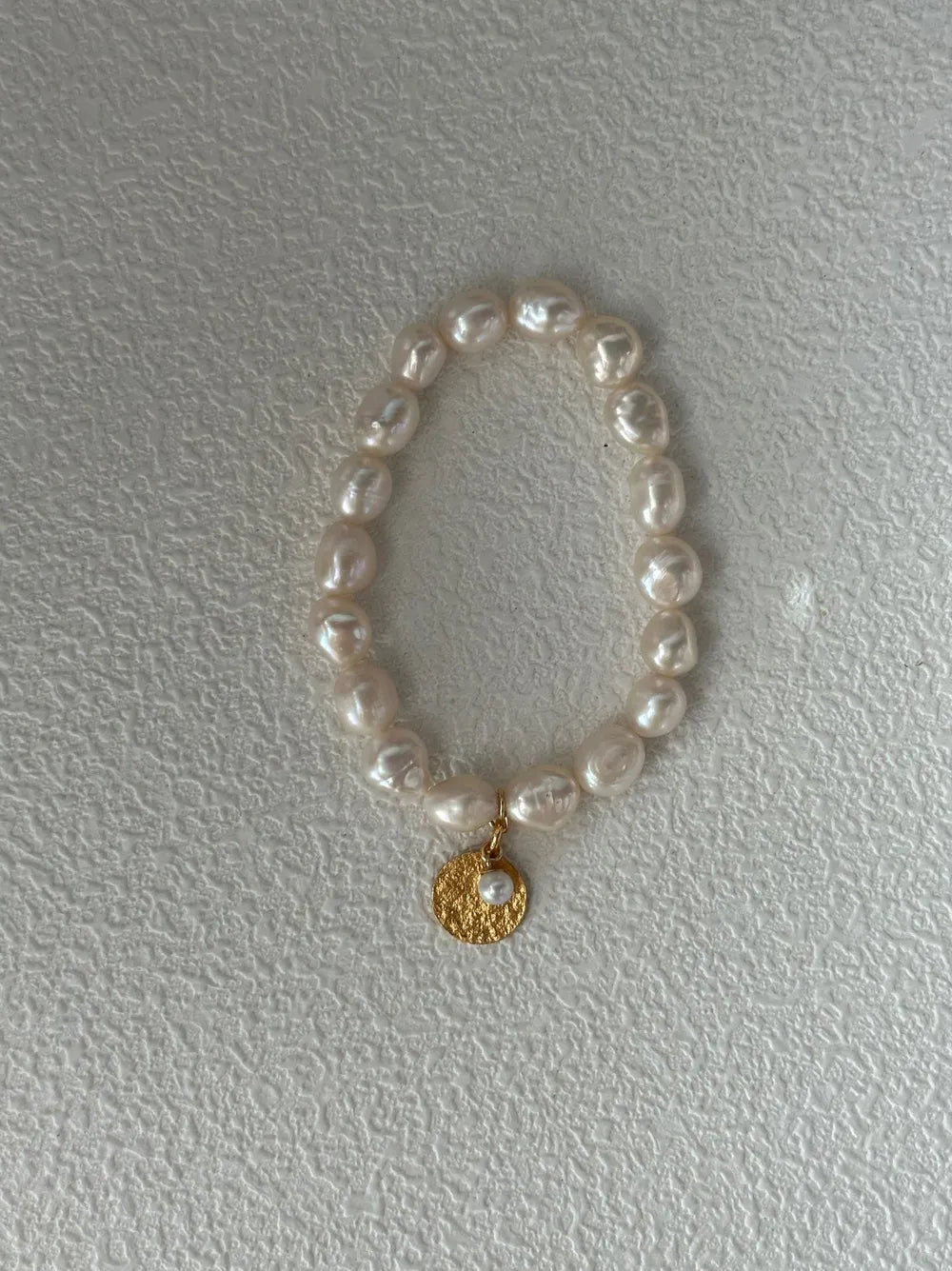 Gardener freshwater pearl elastic bracelet with gold coin charm on white textured backdrop.