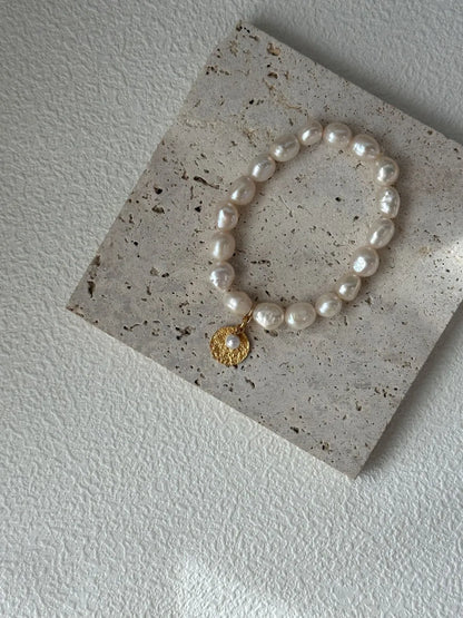 Gardener freshwater pearl elastic bracelet with coin charm resting on square travertine tile.