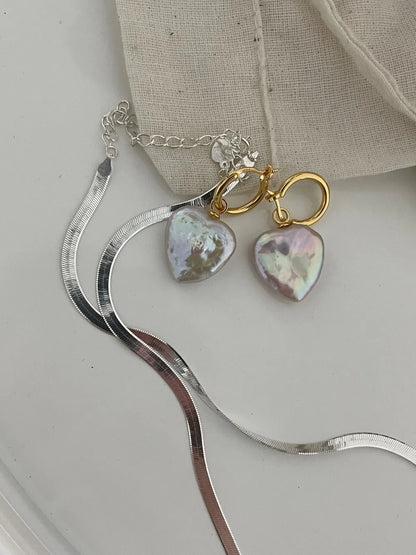 Heart baroque pearl huggie earrings paired with a silver flat herringbone chain on white tabletop.
