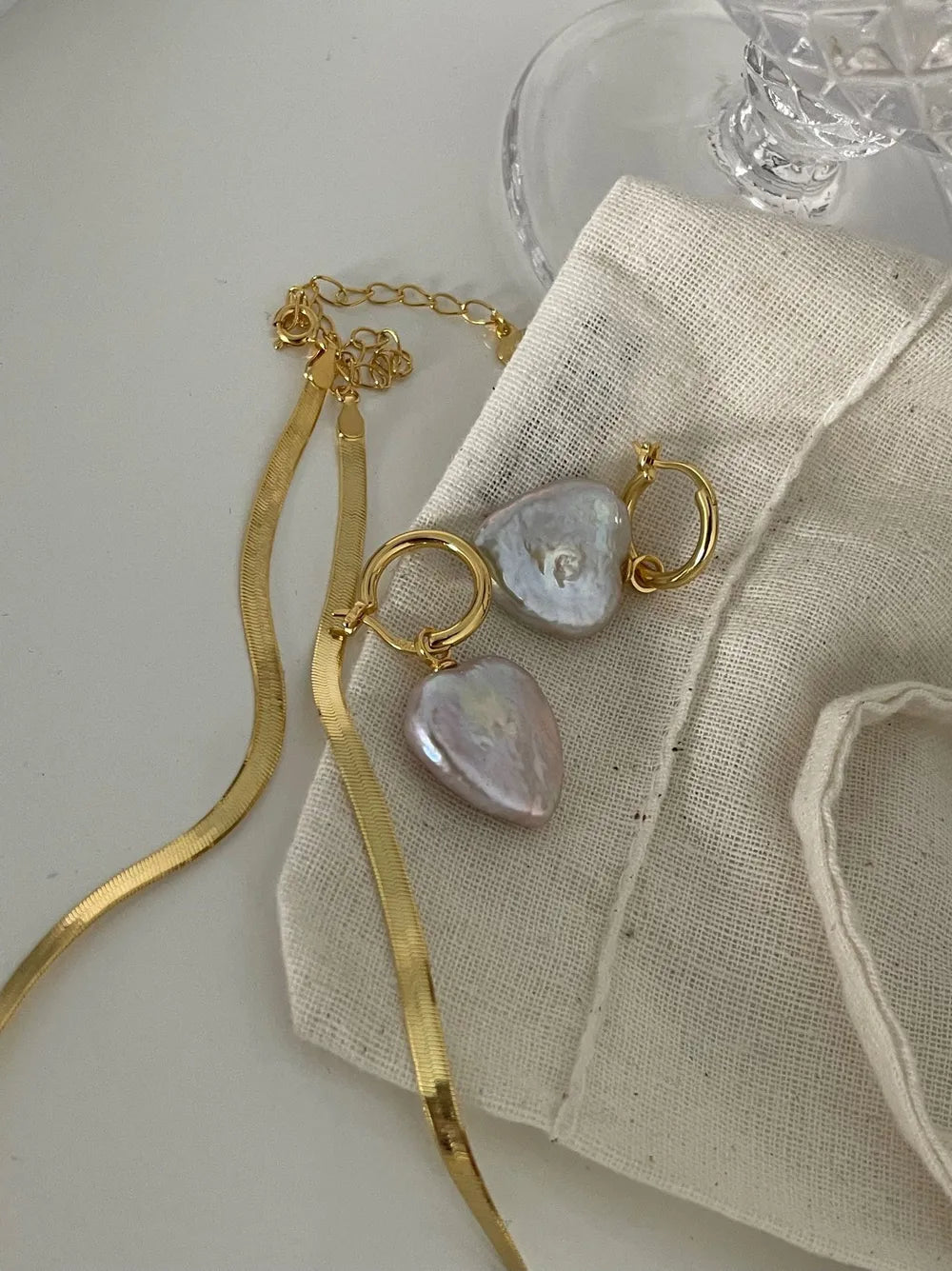 Heart baroque pearl huggie earrings styled with a gold herringbone chain on canvas pouch.