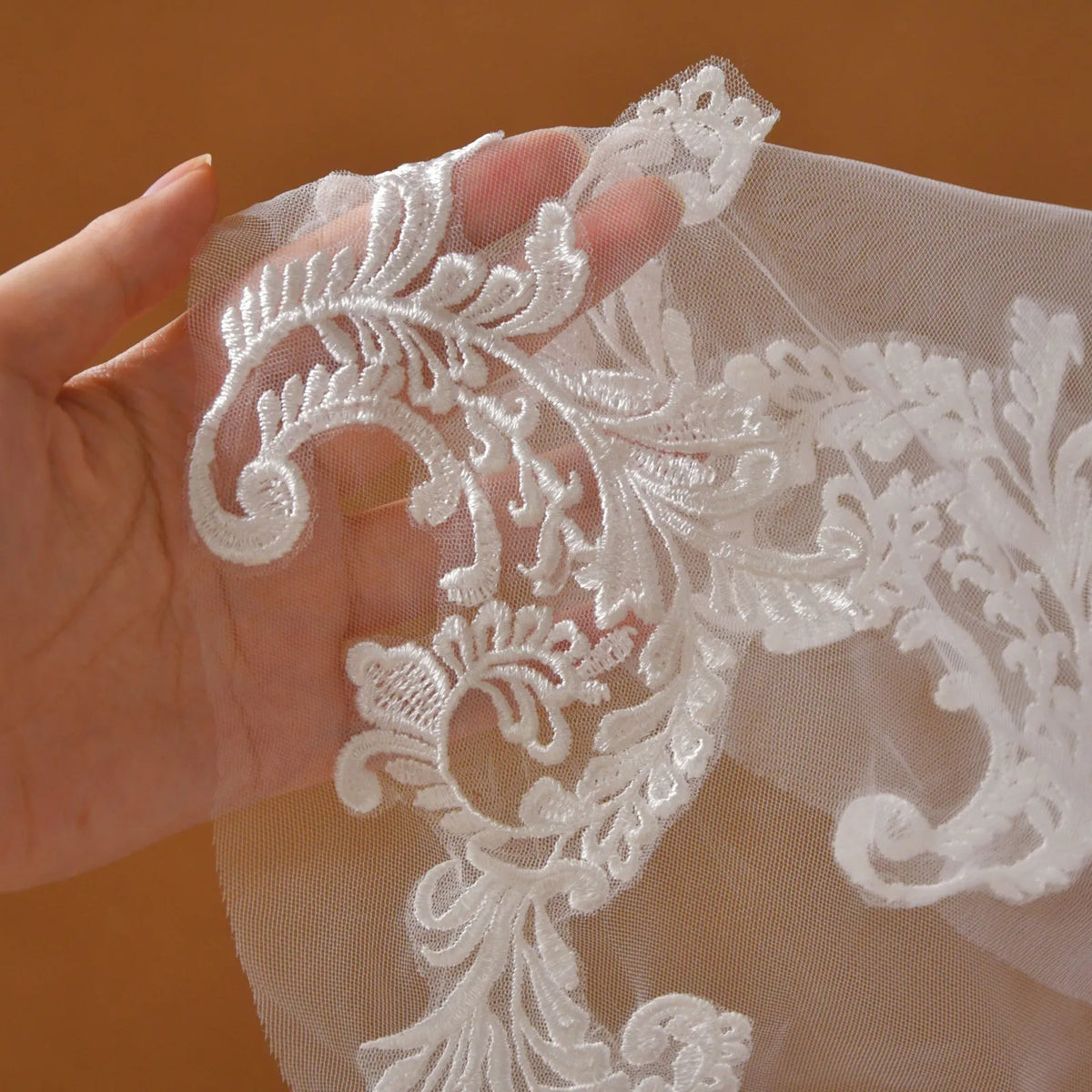 Close-up hand shot showing detailed white embroidered baroque lace appliqué pattern on sheer tulle