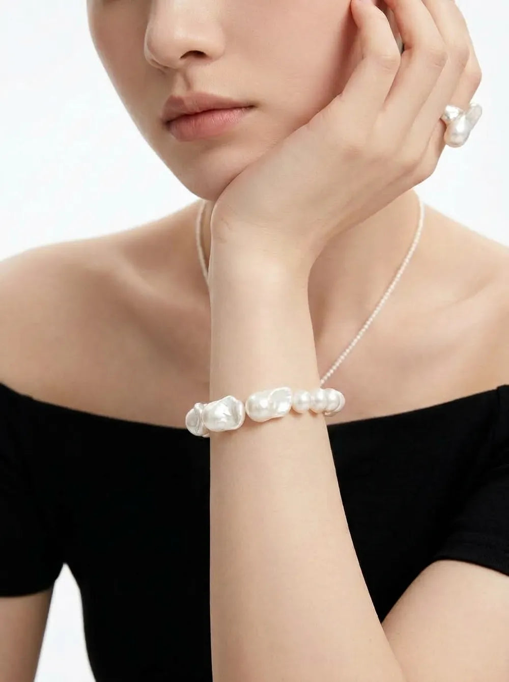 Model in off-shoulder black outfit holding hand to chin showing the large baroque pearl bracelet.