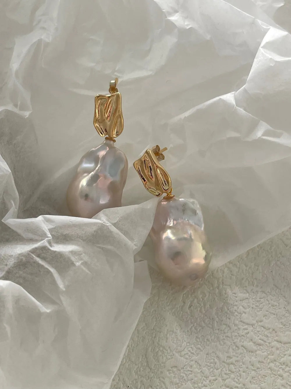 Molten gold metal baroque pearl drop earrings resting on folded white tissue paper.