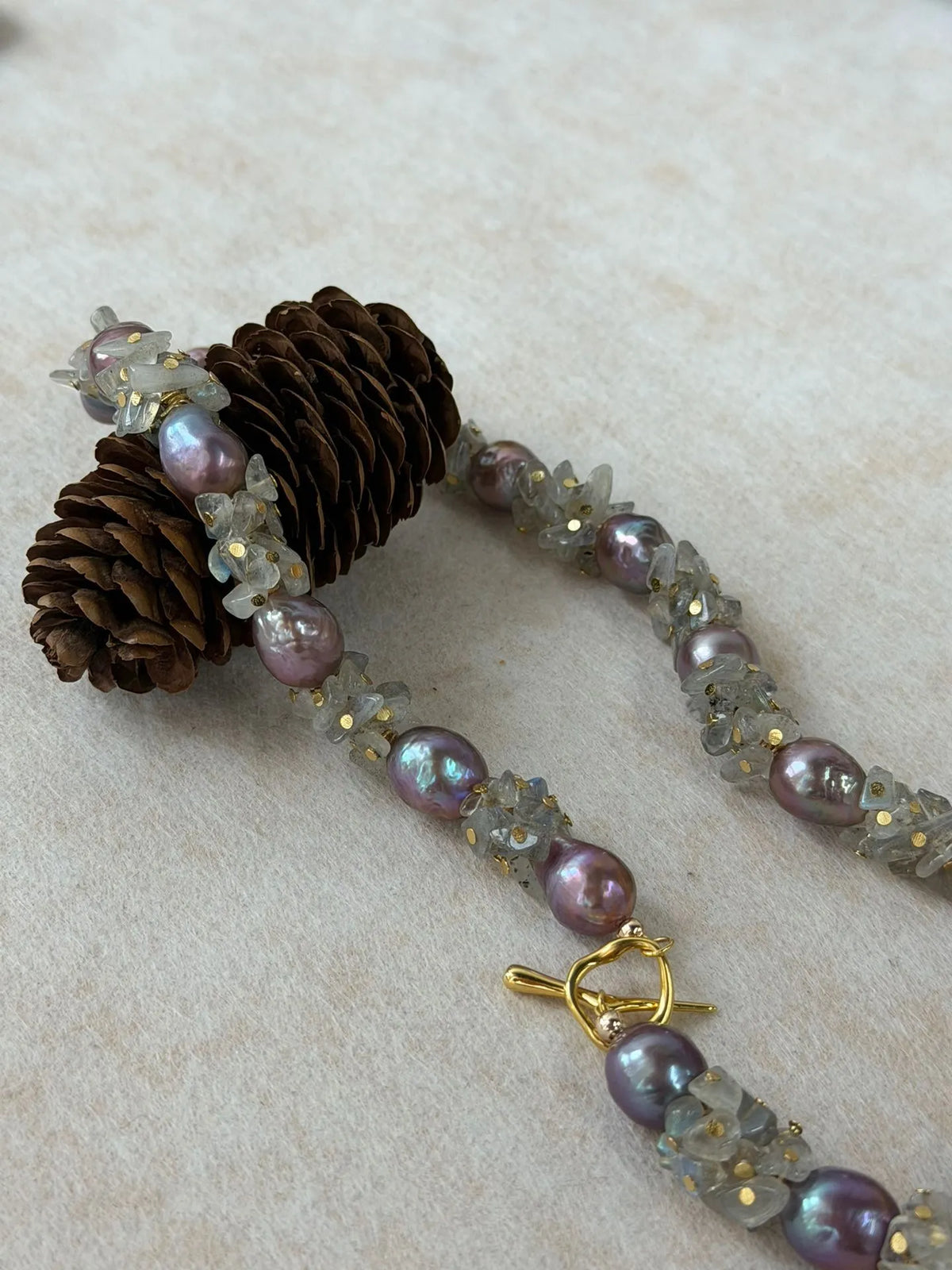 Moonstone and purple baroque pearl necklace draped around a pinecone prop showcasing gold OT clasp.