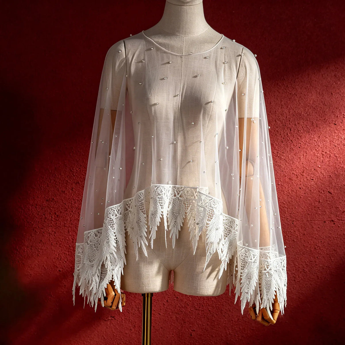 Front view of mannequin displaying sheer pearl bridal capelet against textured red backdrop