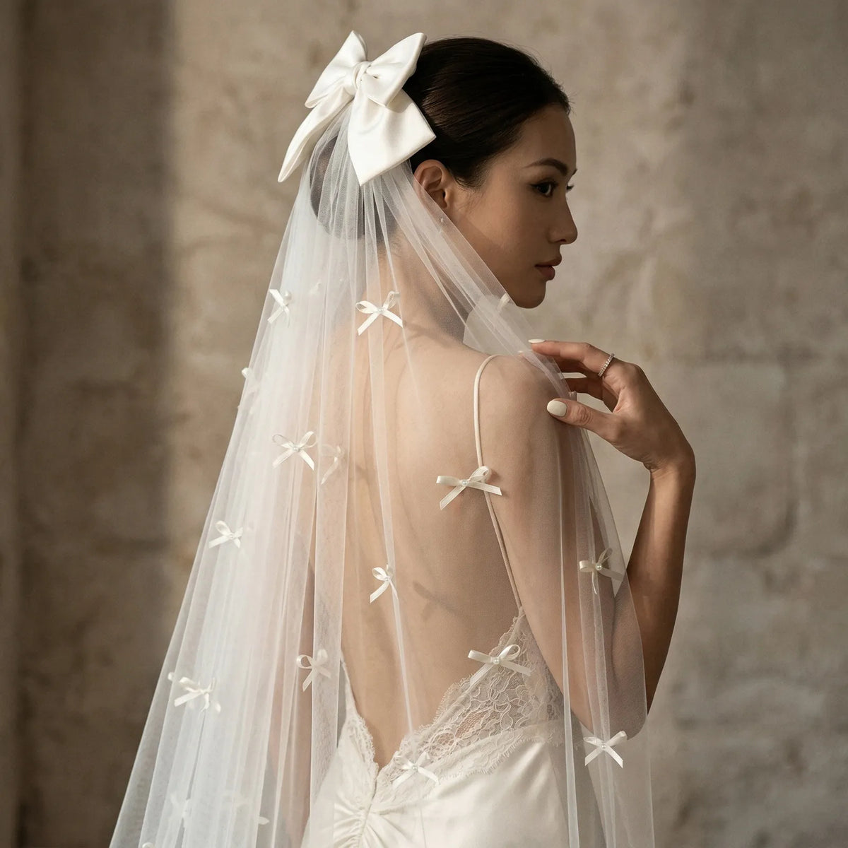 Side view of bride holding veil edge showing scattered pearl-centered ribbon bows and satin top bow