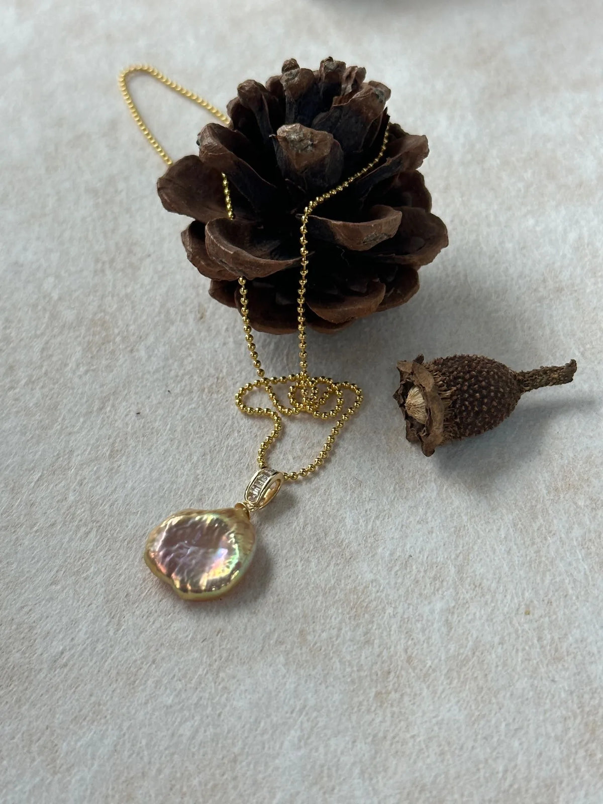 Petal pearl pendant on gold beaded chain displayed with pinecone prop.