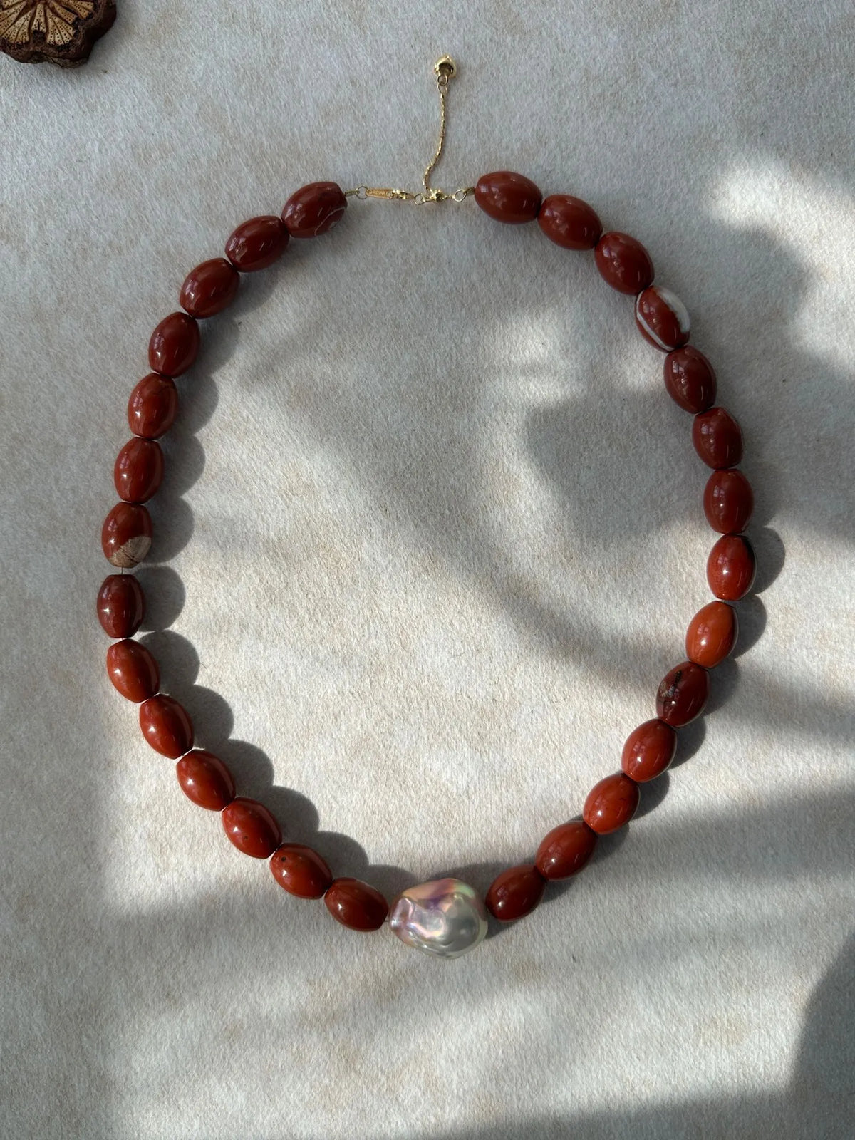 Red jasper beaded necklace with center iridescent baroque pearl under natural sunlight and shadows.
