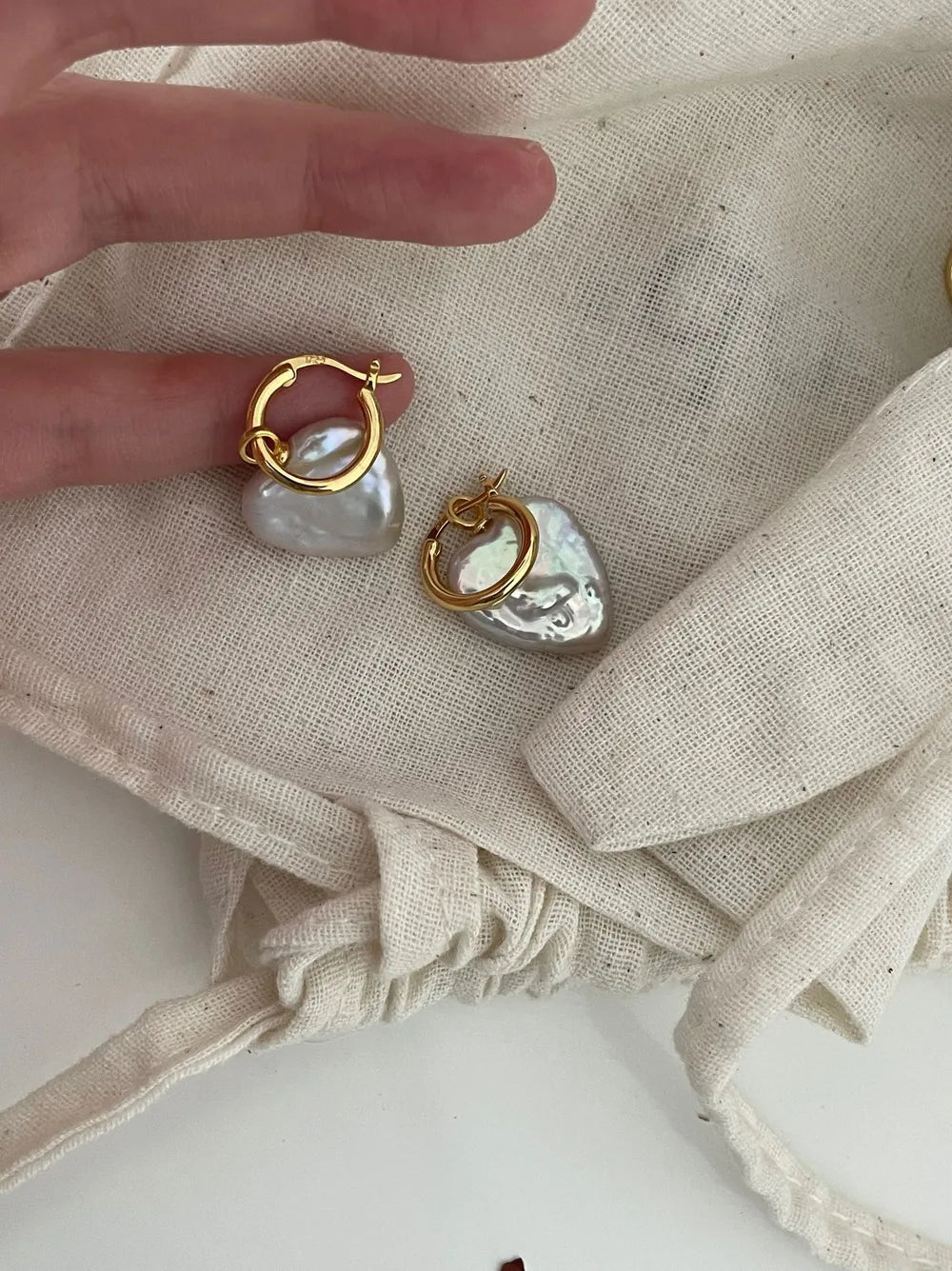 Close-up of hands holding the heart baroque pearl huggie earrings showing S925 hallmark stamp.