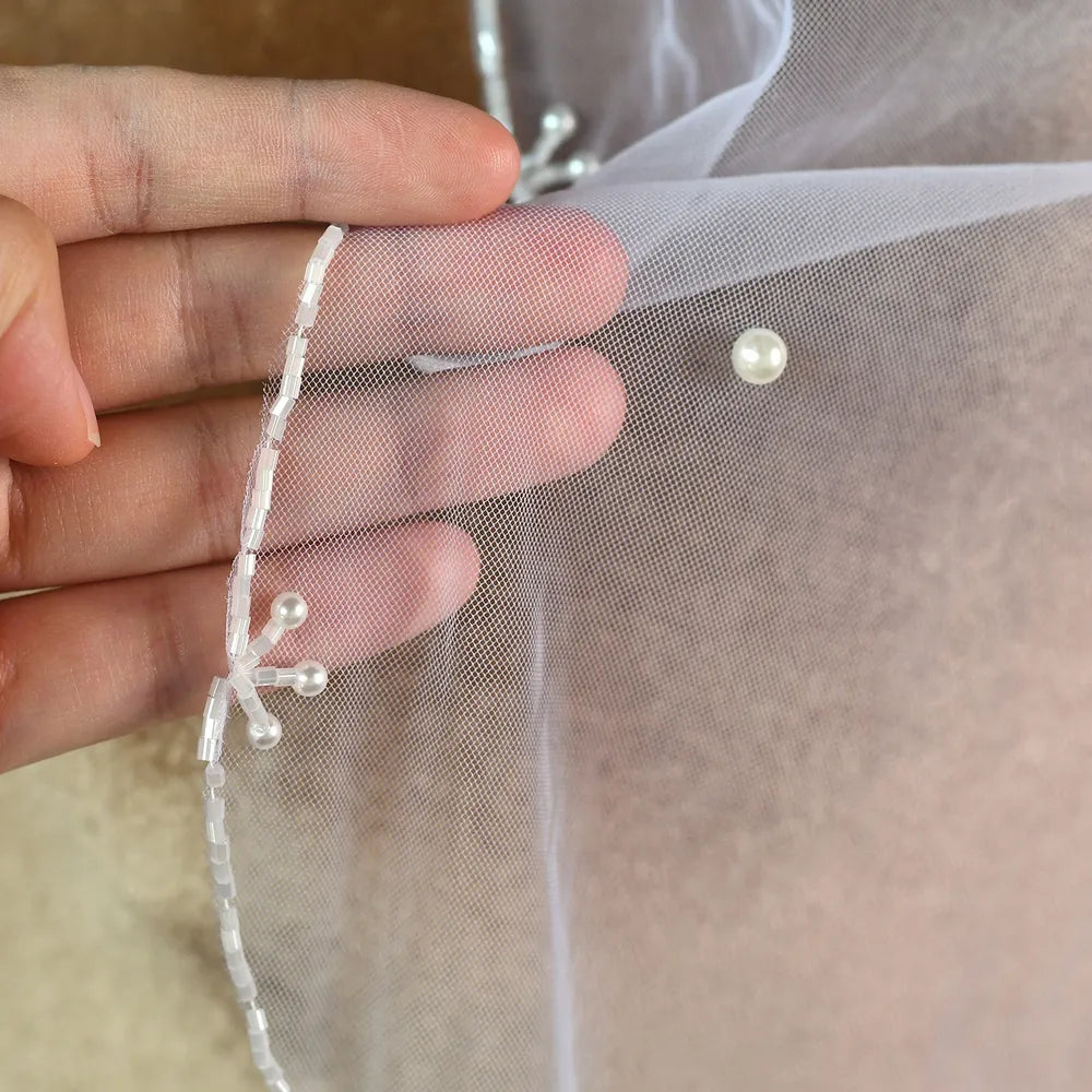 Macro close-up shot showing bugle bead piping, three-pearl floral sprig motifs, and scattered pearls on fine mesh tulle
