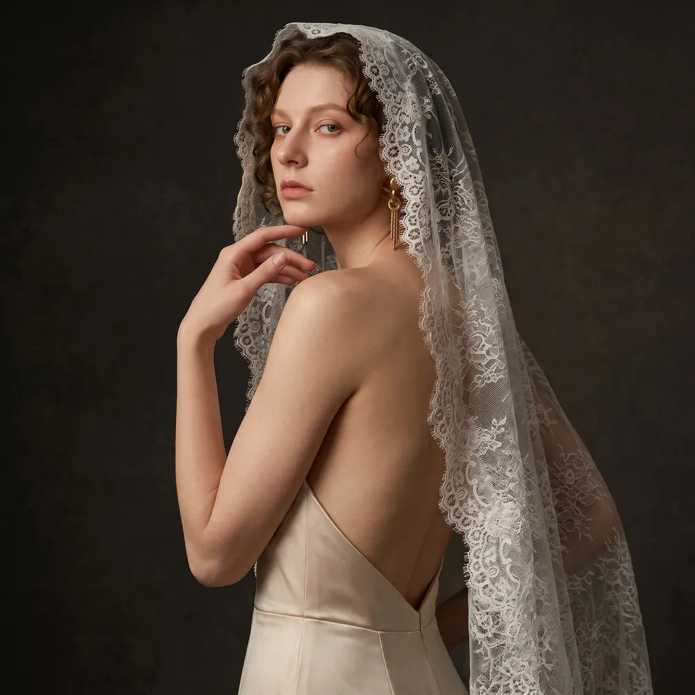 Side portrait of bride in a backless satin gown looking over her shoulder, wrapped gracefully in a sheer lace mantilla veil