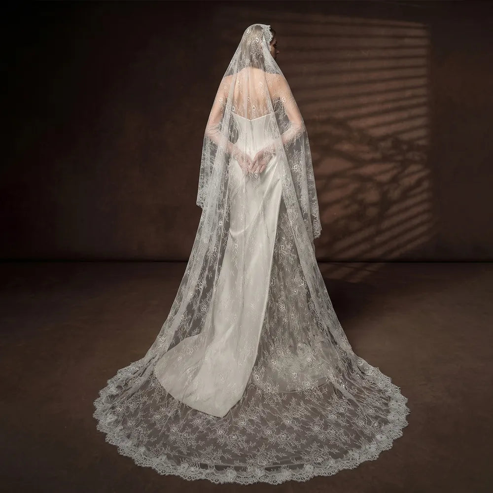 Full-length portrait of bride in a sleek white gown standing in a sunlit garden, wearing a 300cm cathedral lace veil extending onto the stone patio