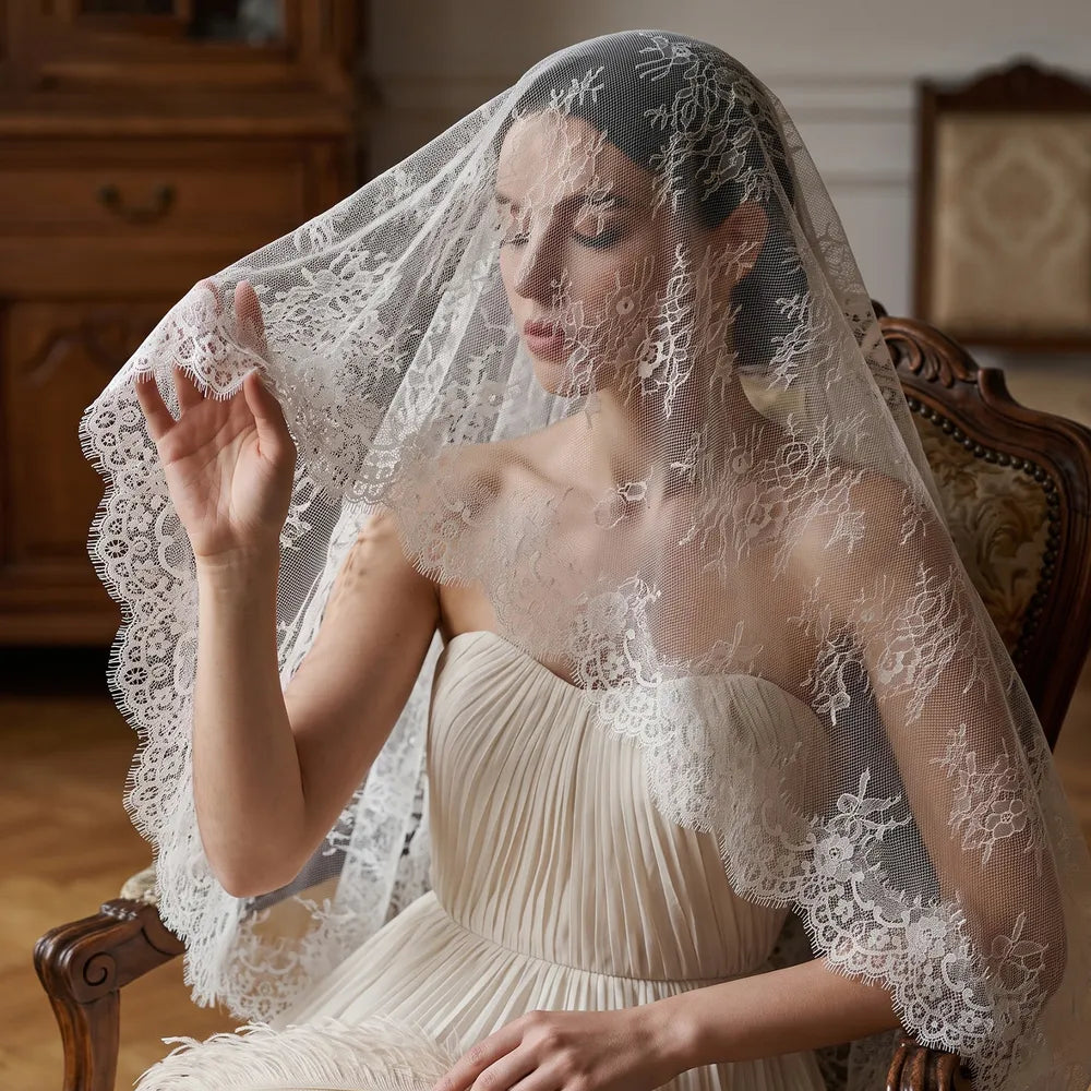 Bride in pleated strapless bridal dress sitting on a vintage wooden armchair, draped softly in a sheer white lace mantilla veil