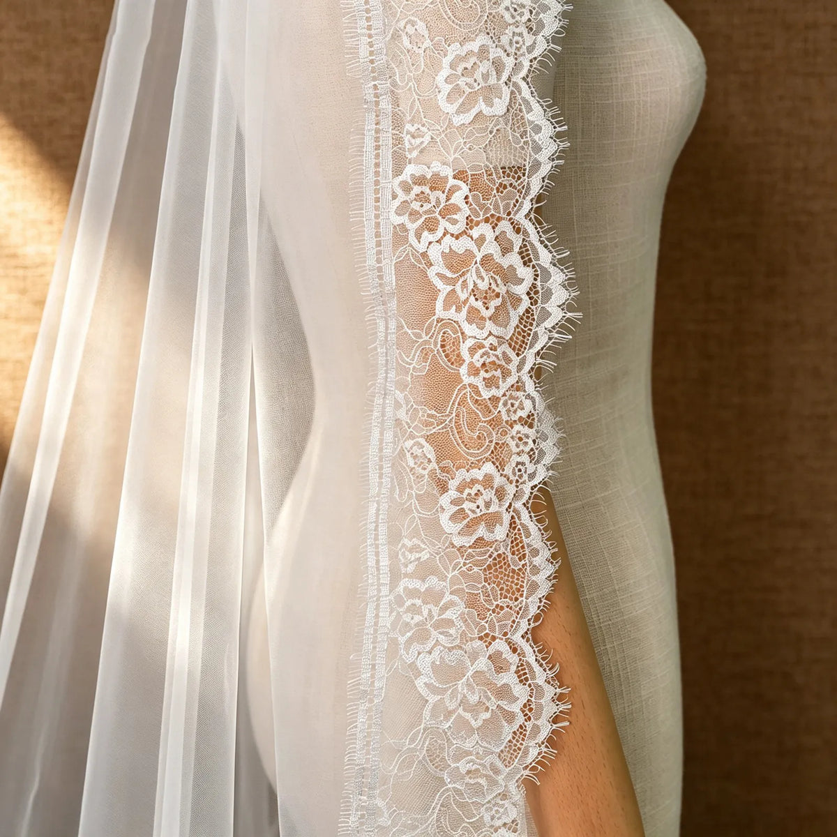 Close-up side profile of the veil on a mannequin showing soft white tulle folds alongside rich floral eyelash lace edging against a warm textured backdrop