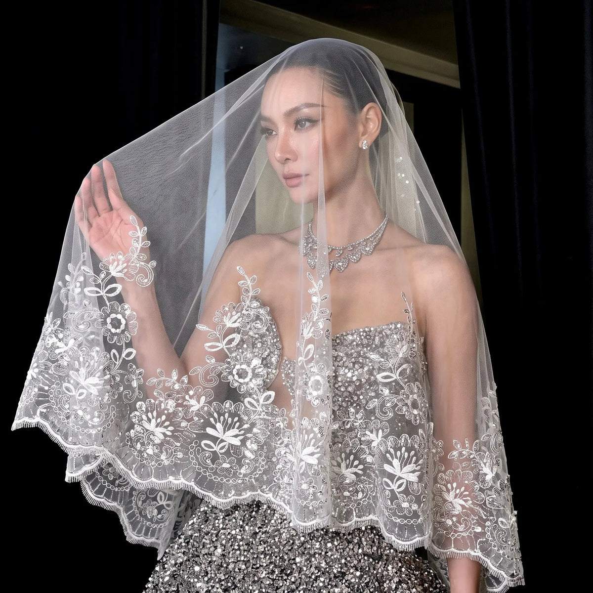 High fashion shot of bride holding sheer blusher veil up, showing heavy sequin lace embroidery and sequined gown