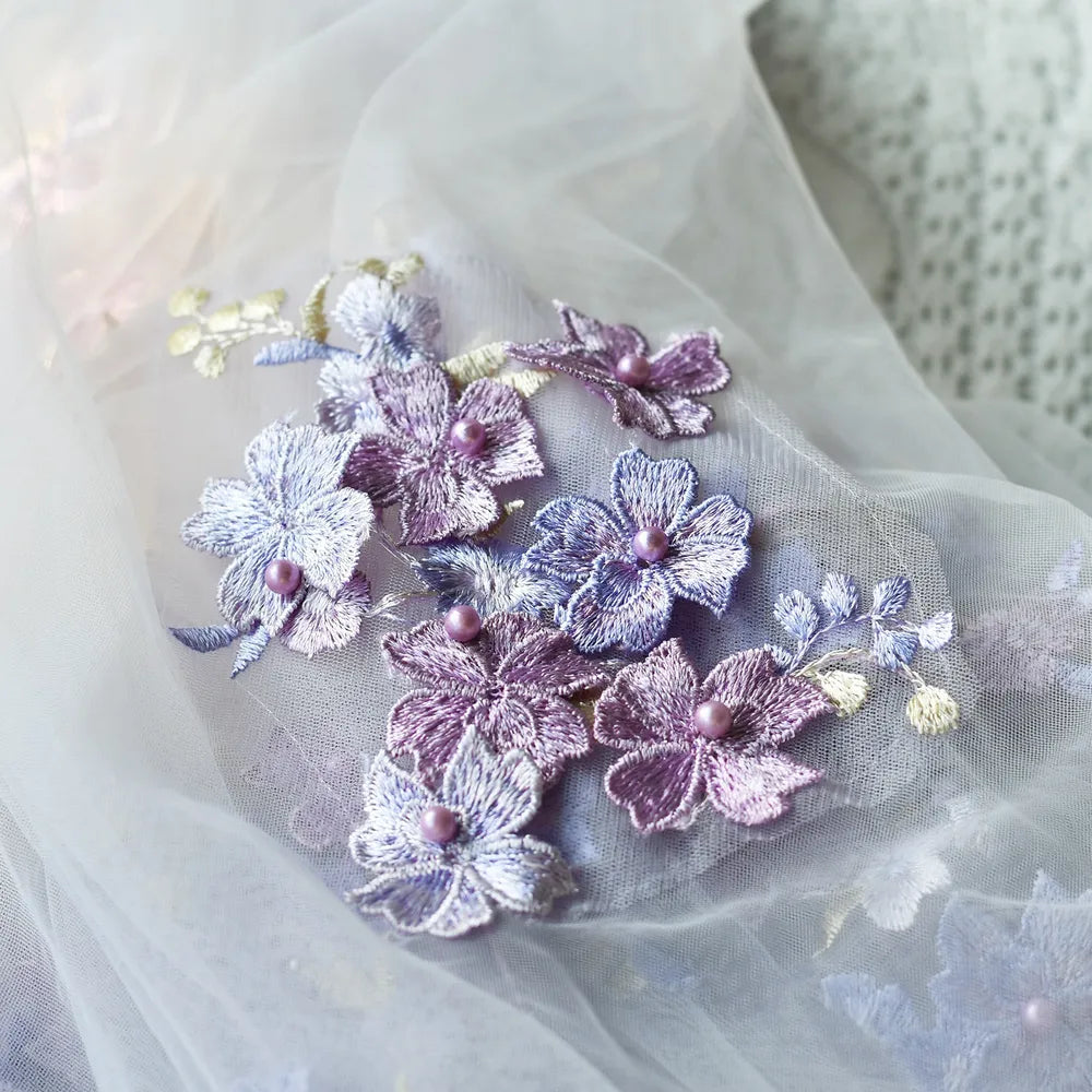 Macro photography close-up highlighting 3D lavender, lilac, and purple embroidered flower appliques with hand-set central pearls on soft white bridal tulle fabric