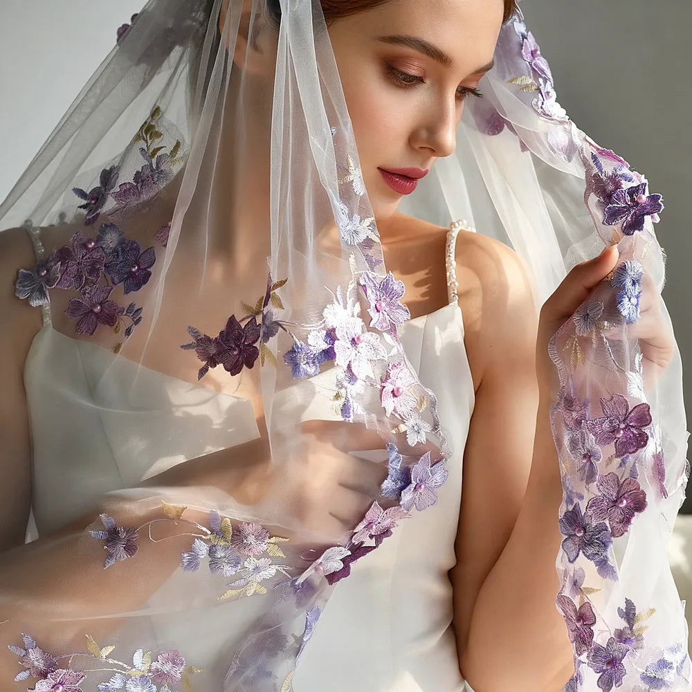 Close-up portrait of bride holding the edge of sheer tulle veil, displaying intricate lilac and dark violet embroidered flowers centered with shimmering pearl beads under natural light