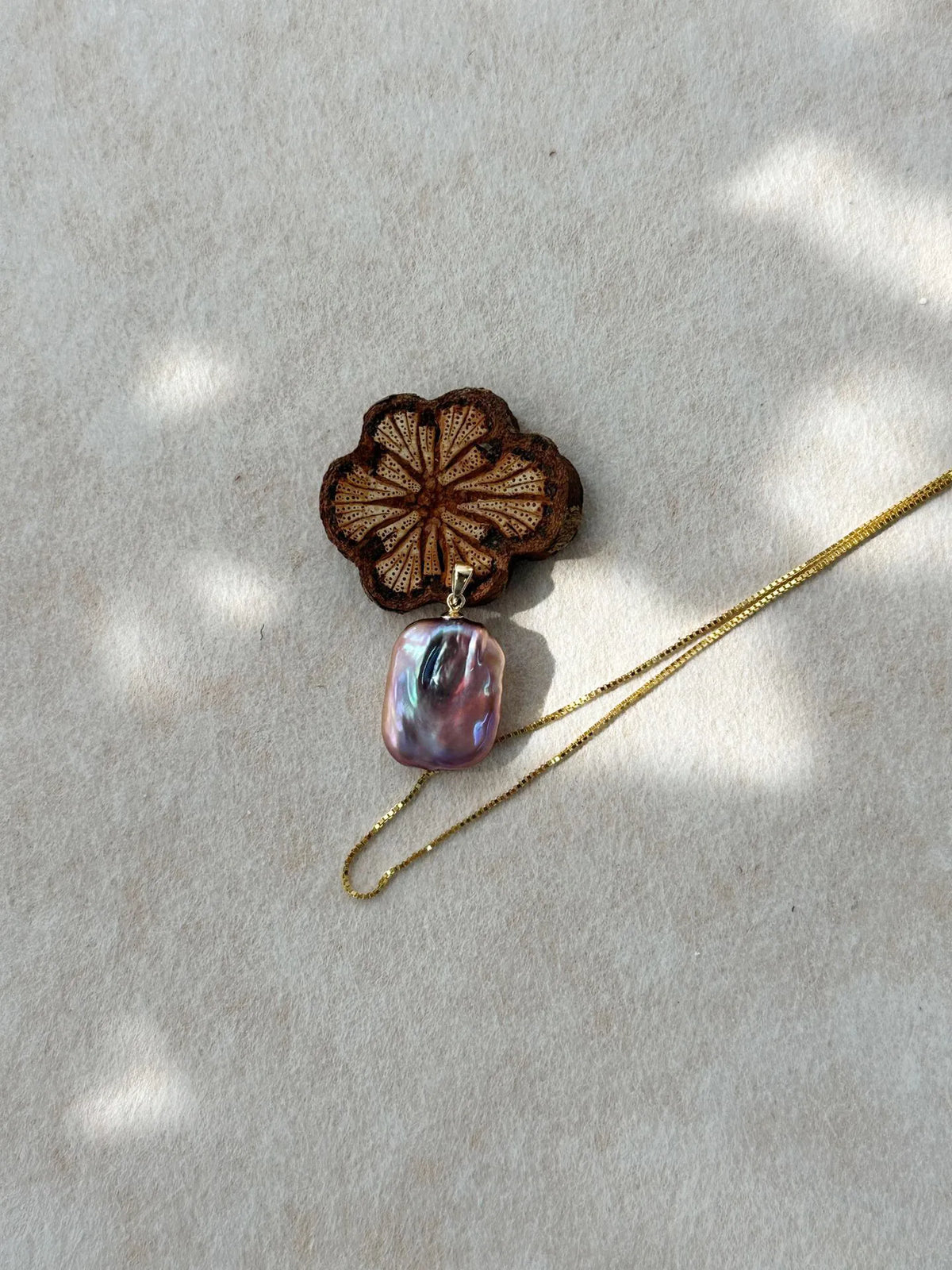 Square baroque pearl pendant displayed next to carved wooden prop under soft light shadow.