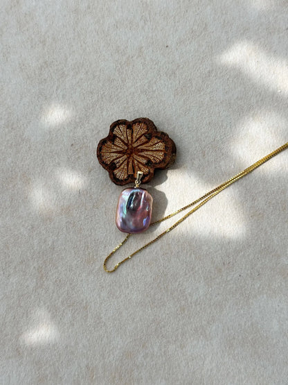 Square baroque pearl pendant displayed next to carved wooden prop under soft light shadow.