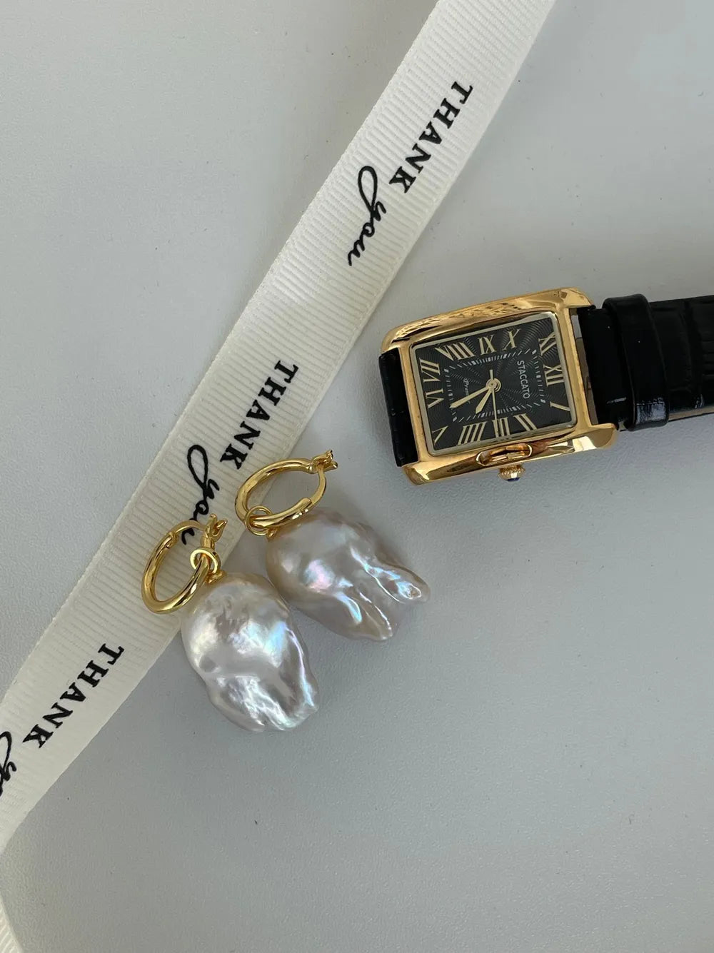Tailed baroque pearl huggie earrings alongside a vintage gold square watch on white surface