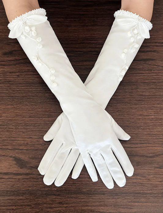 Detailed close-up of white stretch satin bridal gloves with hand-stitched 3D Lily of the Valley organza blossoms, beaded vine stems, and pleated ruffle cuffs.