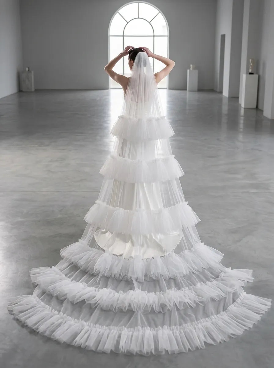 Full length back view of bride in wide studio hall displaying long cascading tiered ruffle veil train