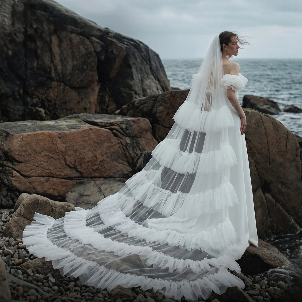 Bride standing on rocky coast showing long 300cm tiered ruffle veil spreading across coastal rocks