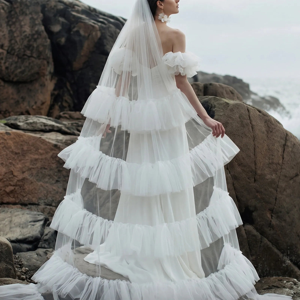 Back view of off-shoulder bridal gown paired with voluminous tiered ruffle tulle veil on rocky beach