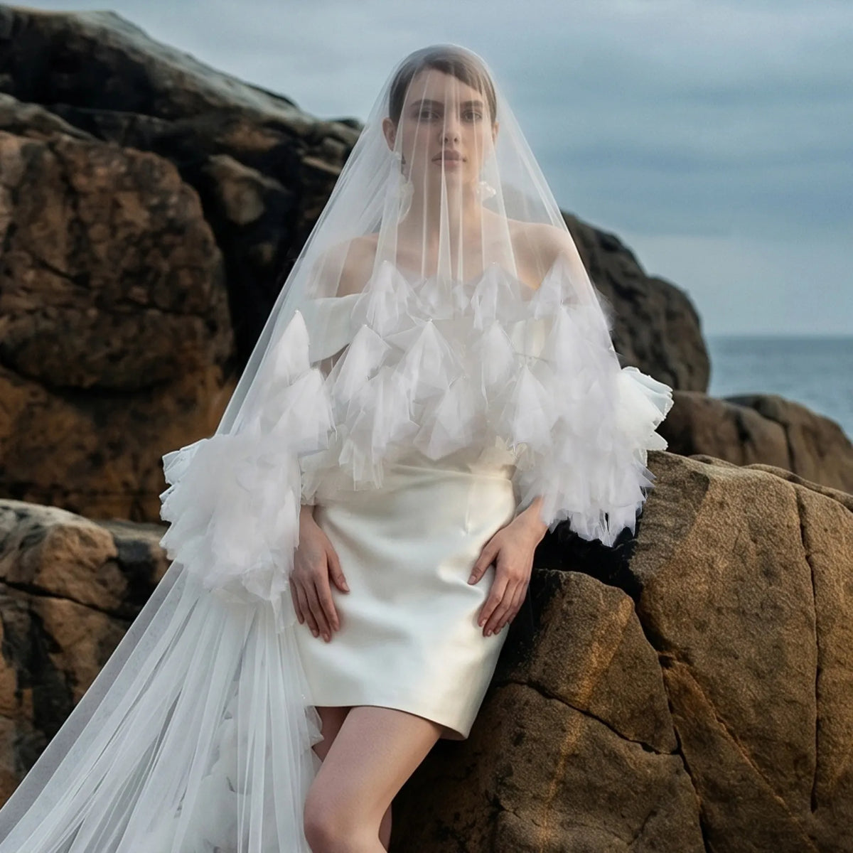 Portrait of bride leaning on sea rocks wearing sheer blusher veil with dramatic ruffled shoulder collar